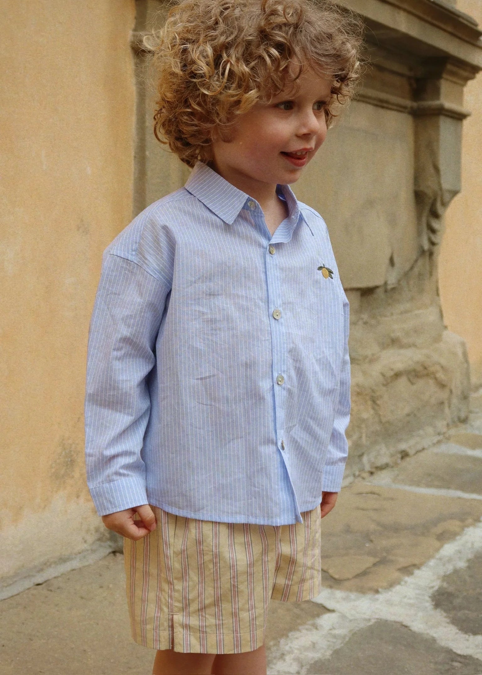 Young boy in a light blue checkered shirt and striped shorts standing against a stone wall.