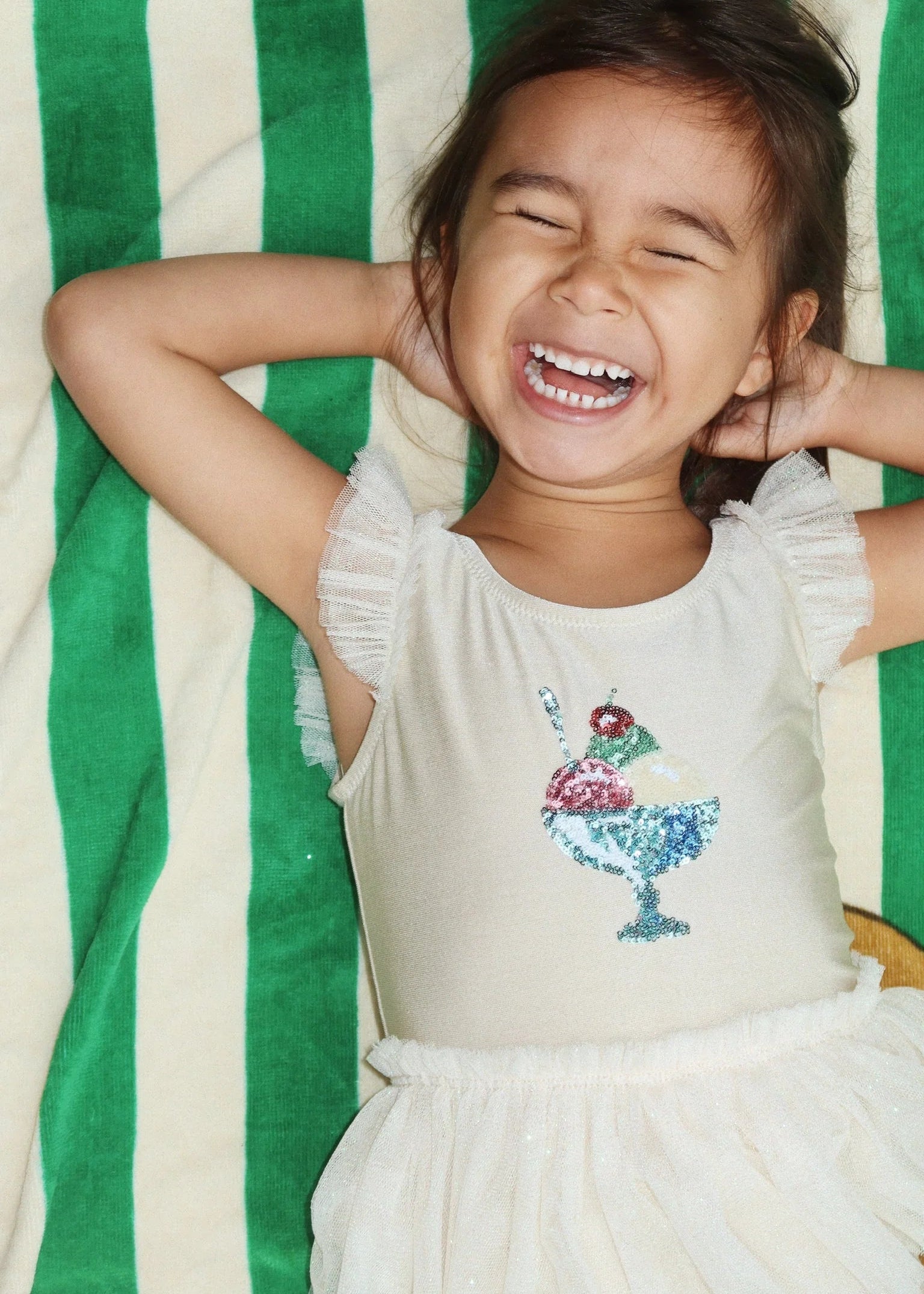 Child wearing a white dress with a colorful design, lying on a green and white striped blanket.