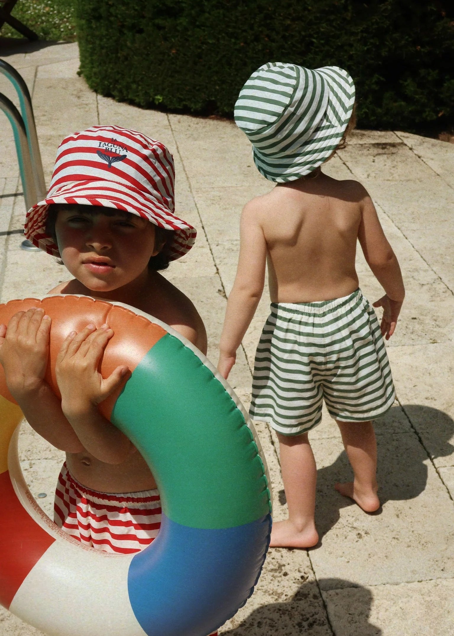 Two children wearing colorful hats and swimwear, one holding a life buoy, on a sunlit patio.