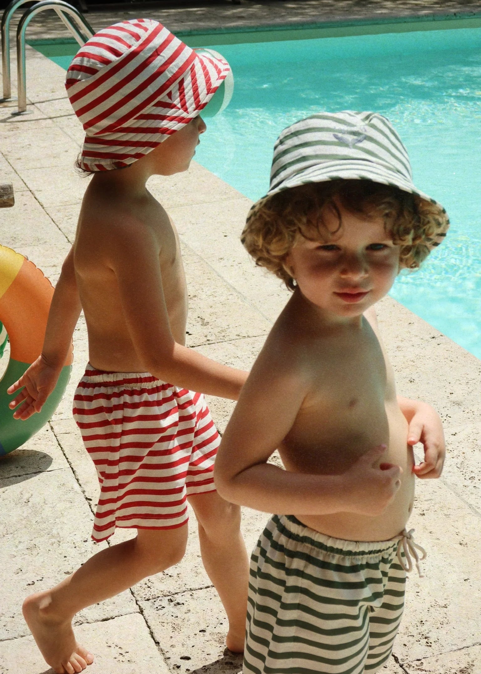 Two children in swim hats and striped swimsuits by a pool.
