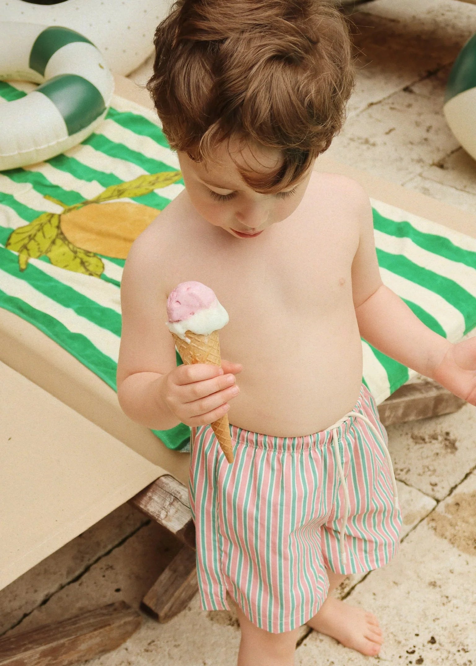 Child holding an ice cream cone on a sandy surface with a striped towel and pool float in the background.