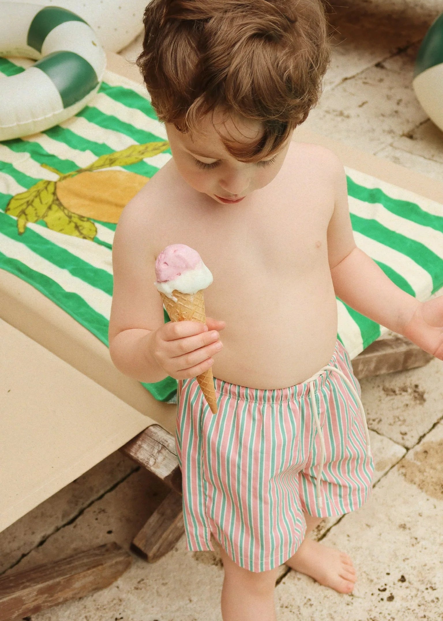 Child holding an ice cream cone on a sandy surface with a striped towel and pool float in the background.