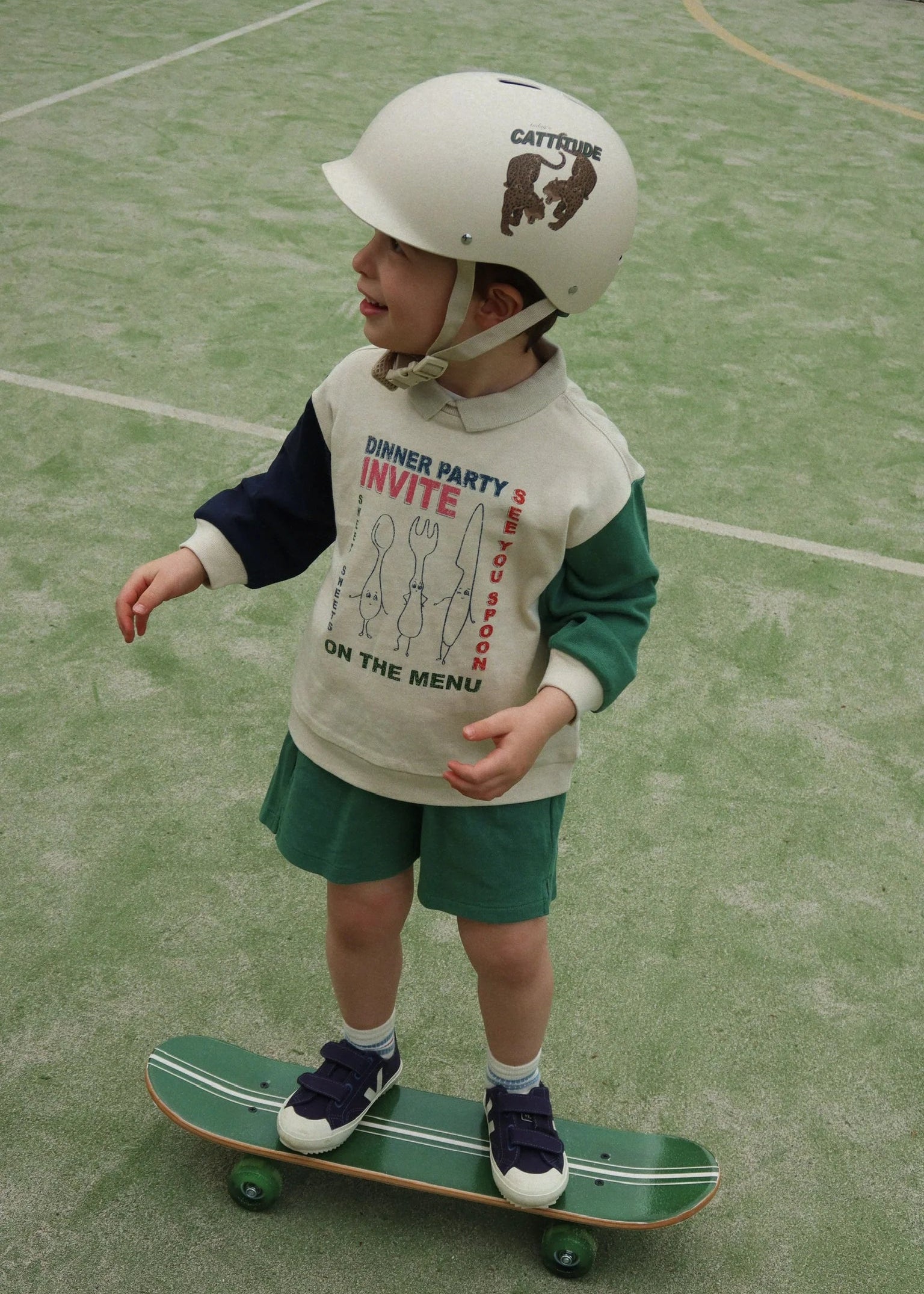 Child wearing a helmet and a sweatshirt with text, standing on a skateboard on a green surface.