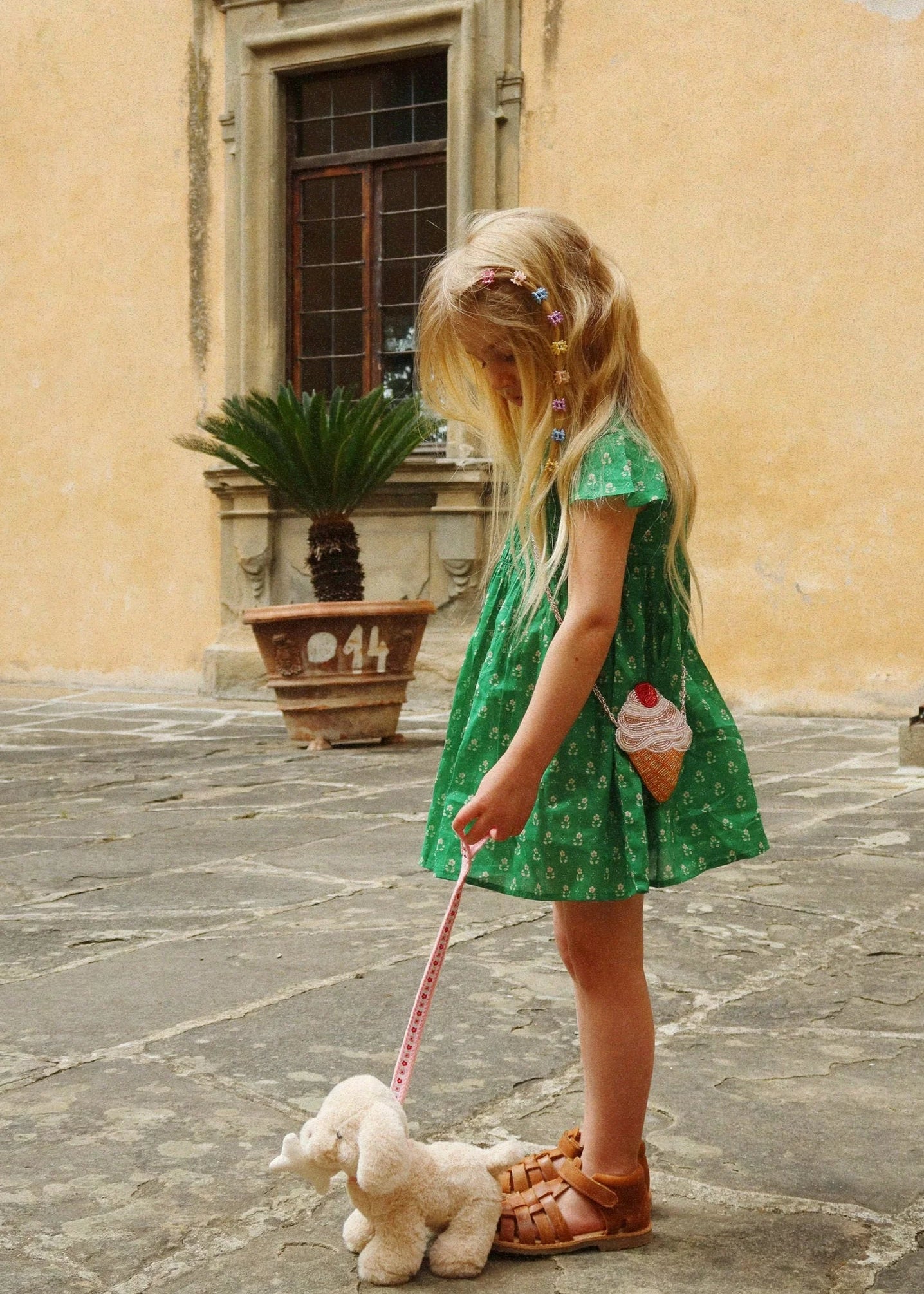 Young girl in a green dress holding a teddy bear in an outdoor setting with a building in the background.