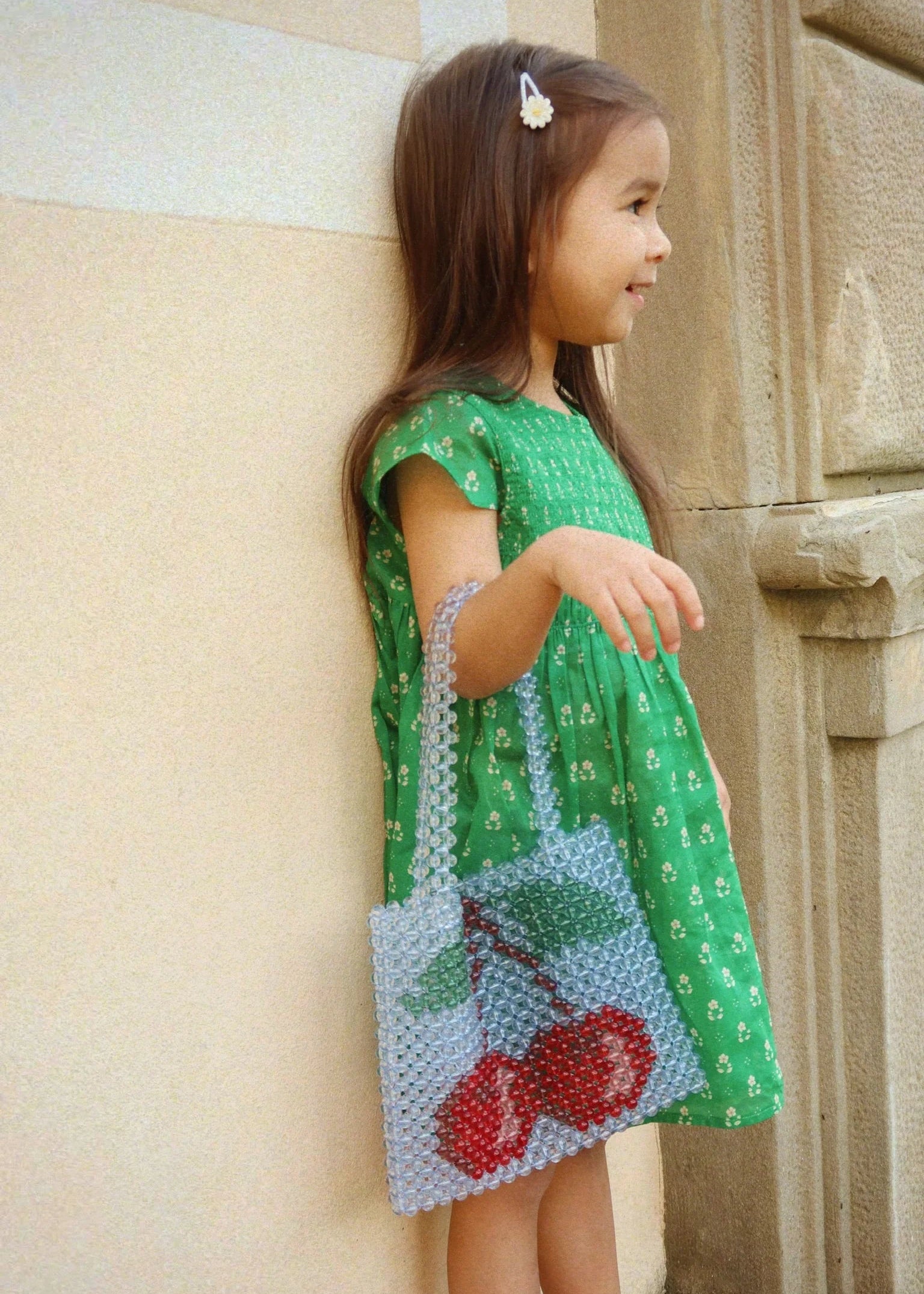 Young girl in a green dress with cherry design leaning against a stone wall.