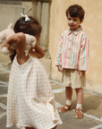Two children, a girl in a white dress holding a teddy bear and a boy in a striped shirt and shorts, standing on a stone pavement.