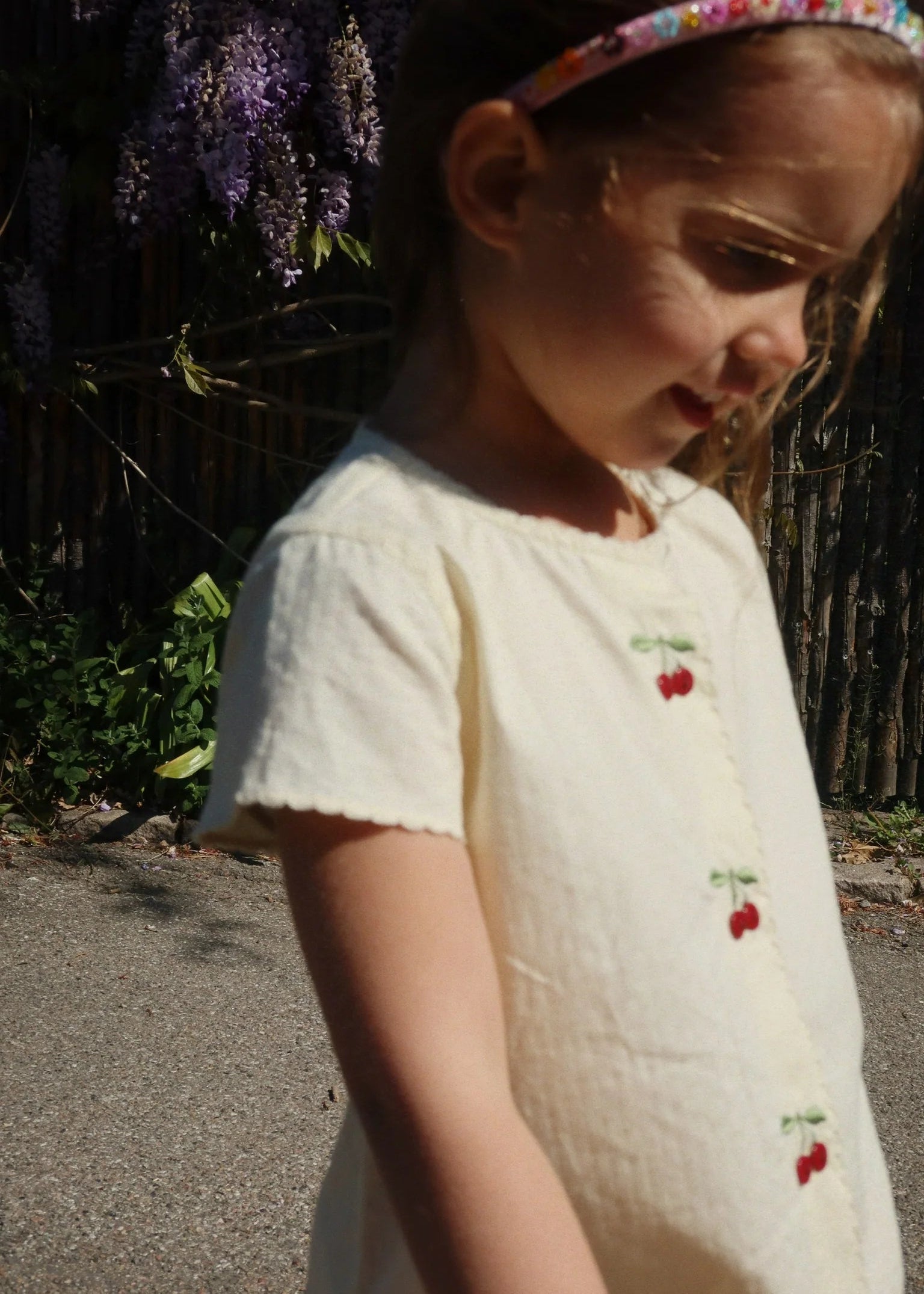 Young girl wearing a white dress with red cherries and a floral headband, standing outdoors.