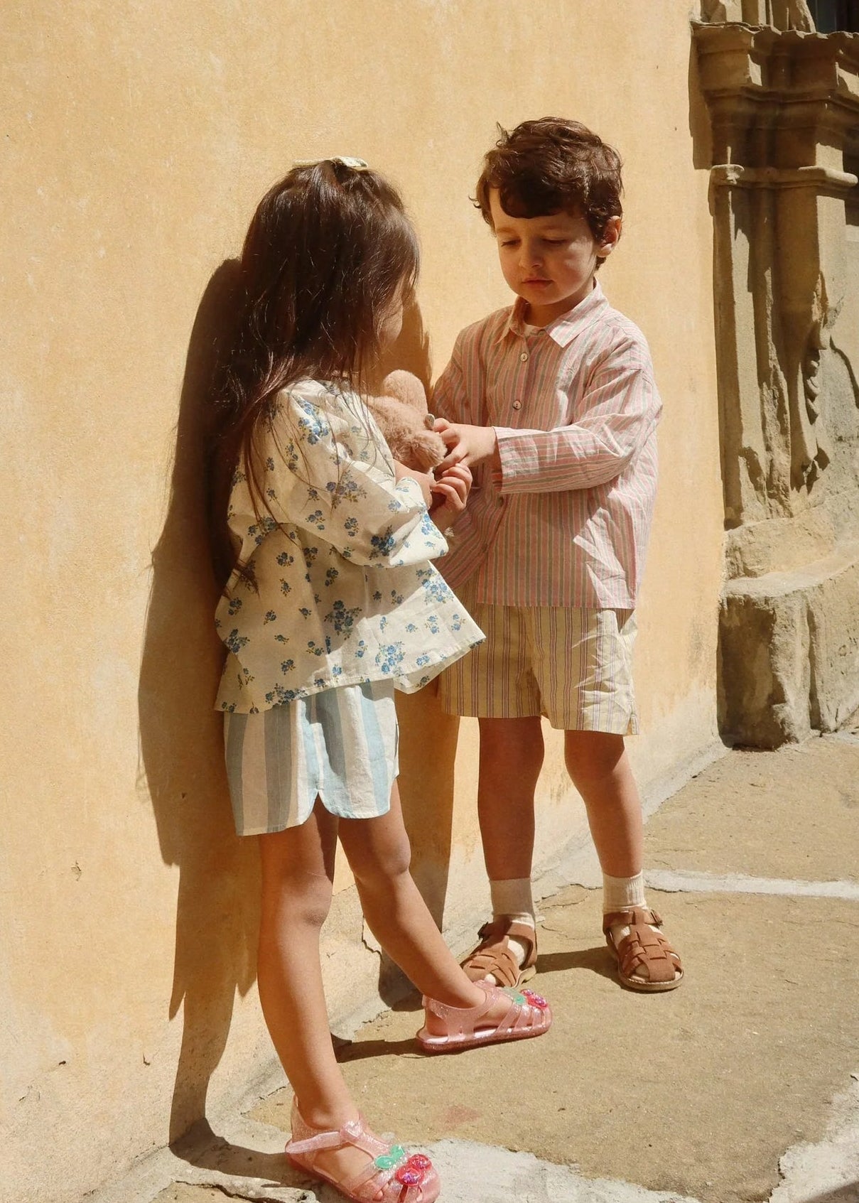 Two children standing close to each other in front of a beige wall.