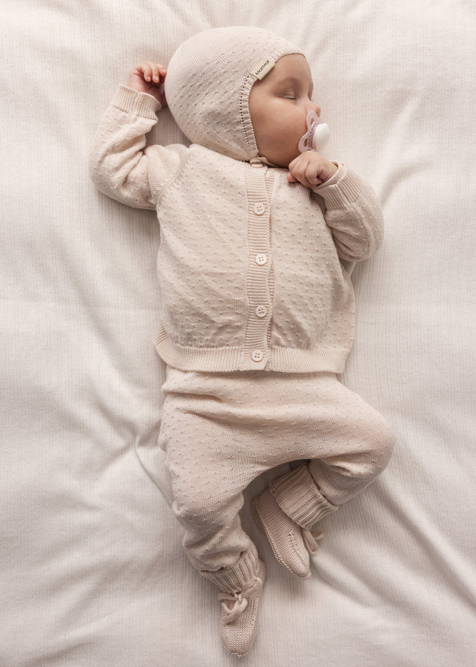 Baby in a cream outfit and hat lying on a white surface