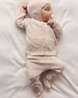 Baby in a cream outfit and hat lying on a white surface