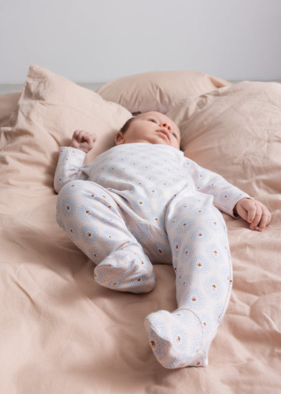 Baby in a white onesie with small patterns lying on a pink bed.