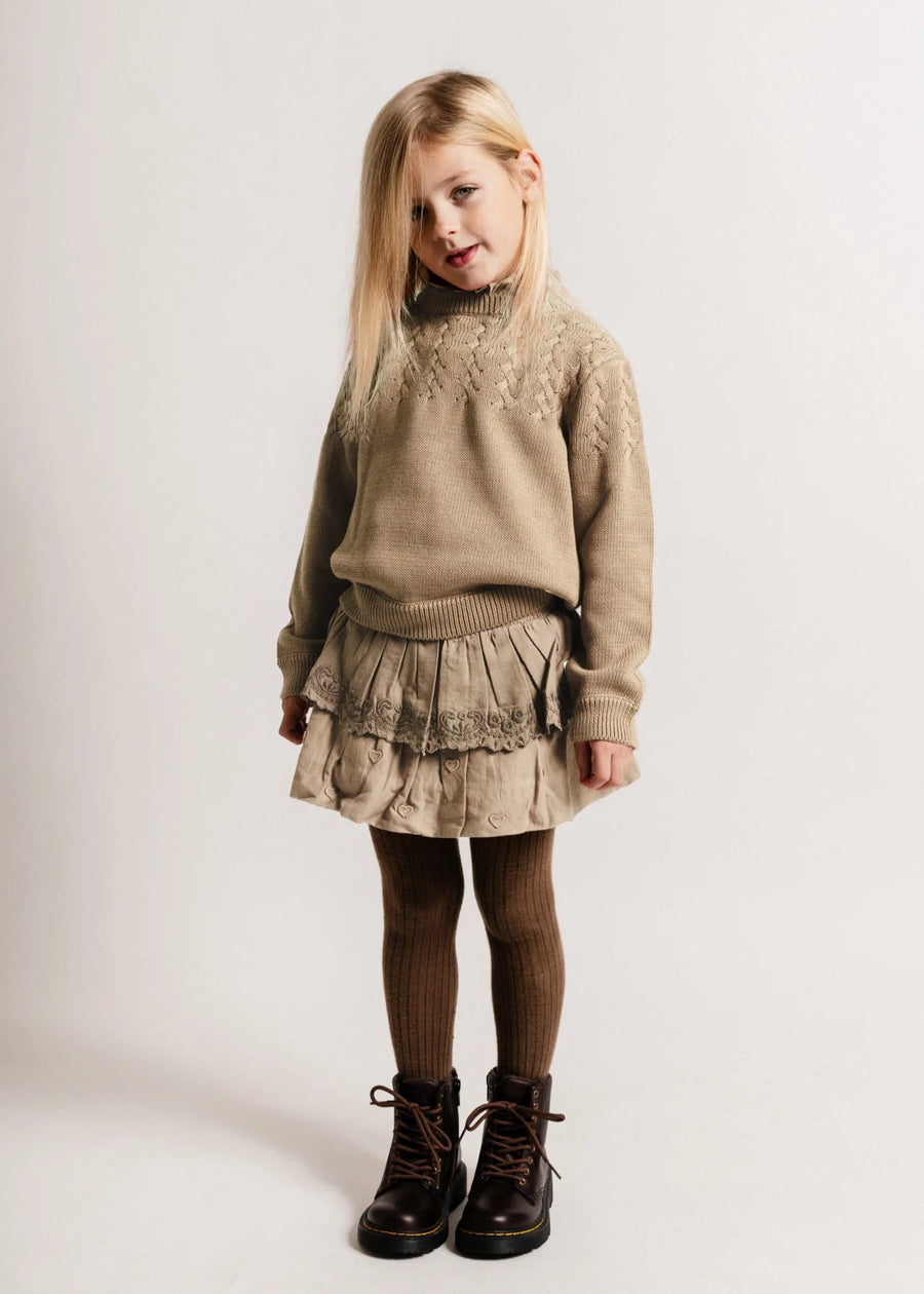 Young girl wearing a beige sweater and skirt with brown boots on a white background