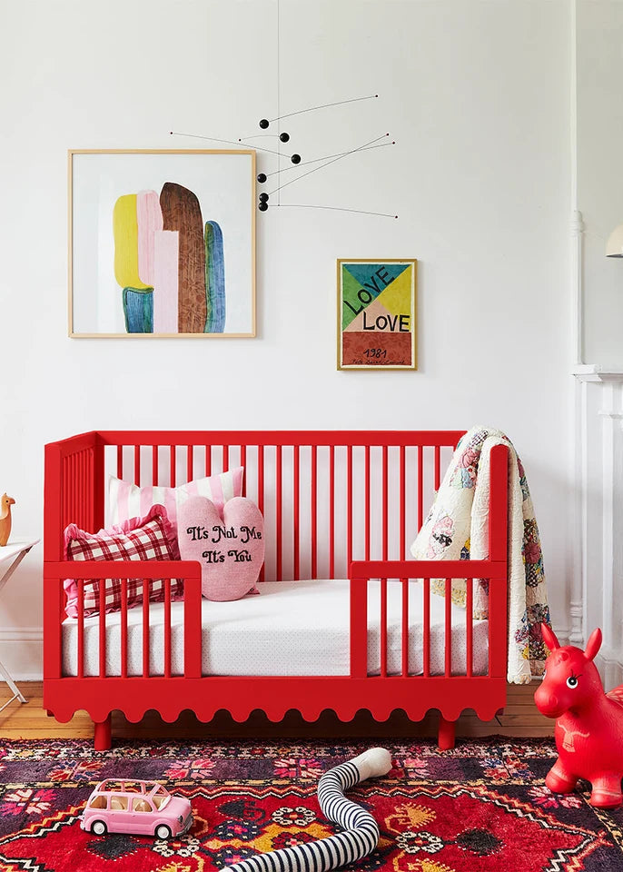 Children's room with a red crib, colorful artwork, and toys on a patterned rug.