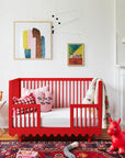 Children's room with a red crib, colorful artwork, and toys on a patterned rug.