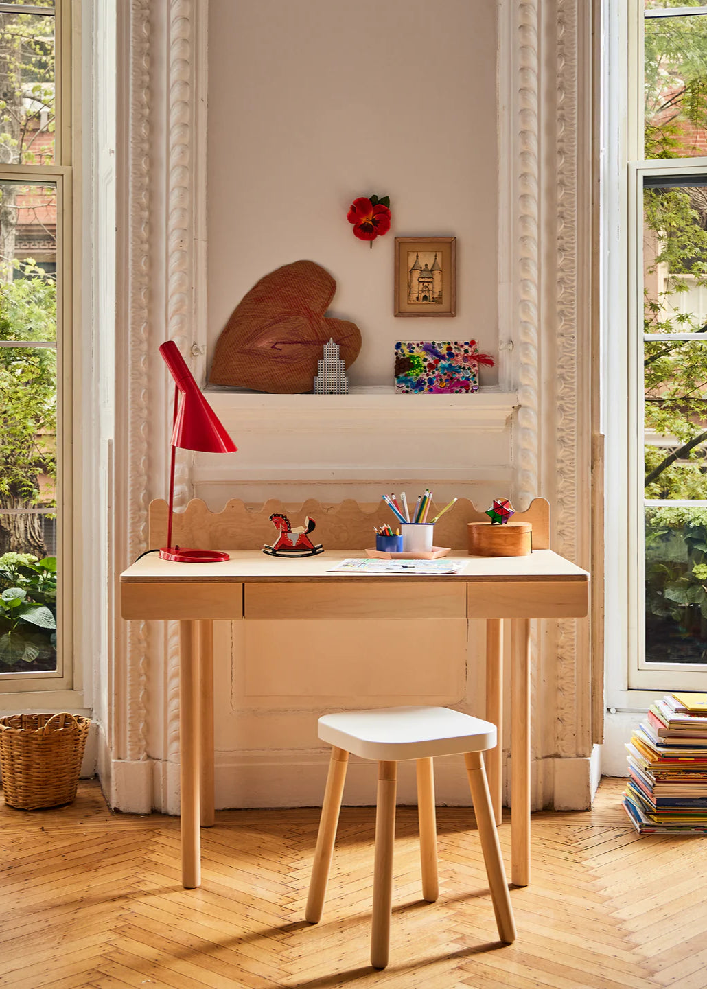 Wooden desk with a red lamp, books, and small decorative items in a room with large windows.