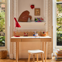 Wooden desk with a red lamp, books, and small decorative items in a room with large windows.