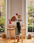Child sitting at a desk in a bright room with large windows, surrounded by books and a red lamp.