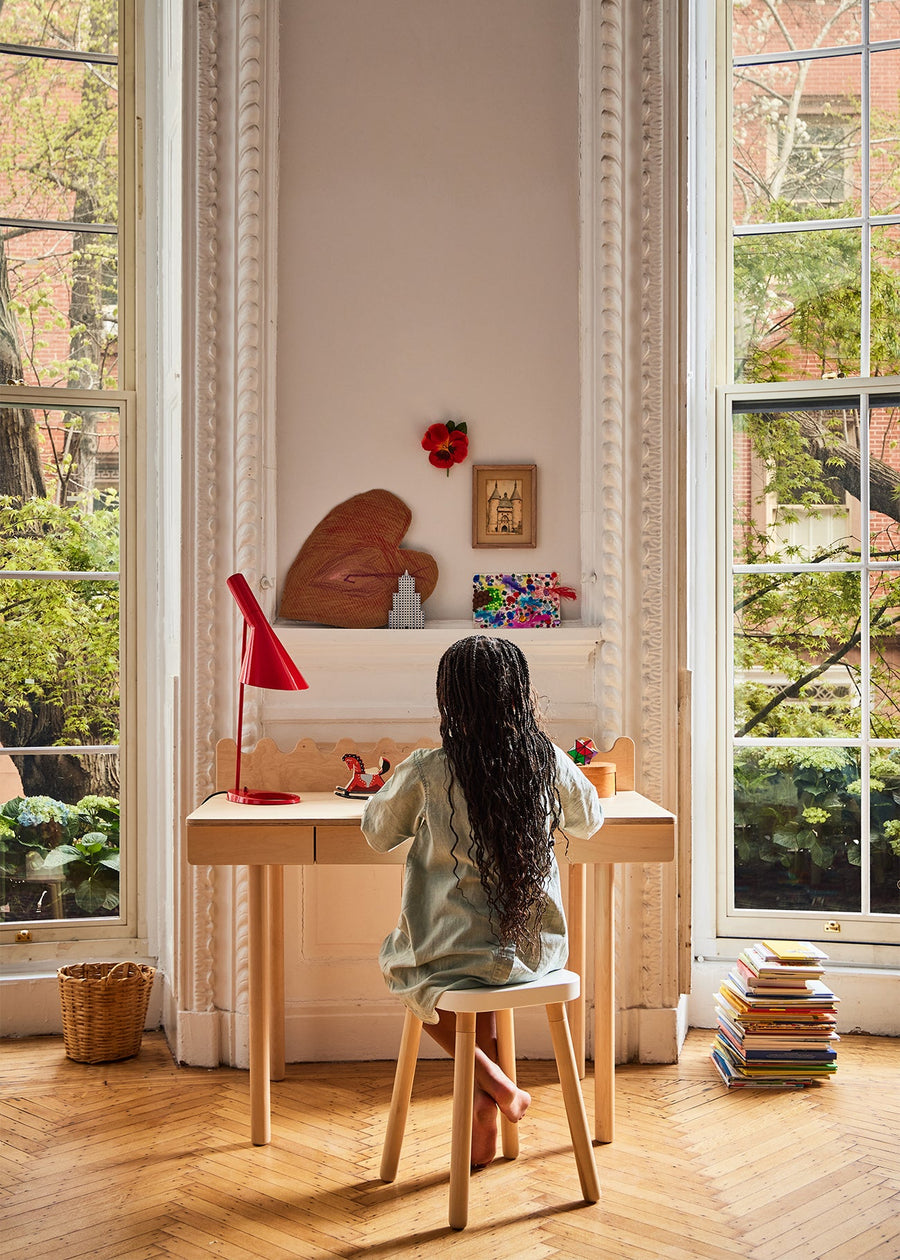 Child sitting at a desk in a bright room with large windows, surrounded by books and a red lamp.