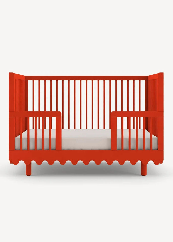 A bright red wooden baby crib with vertical slats and a scalloped bottom edge, shown from the front on a light gray background.
