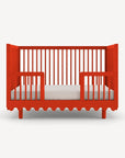 A bright red wooden baby crib with vertical slats and a scalloped bottom edge, shown from the front on a light gray background.