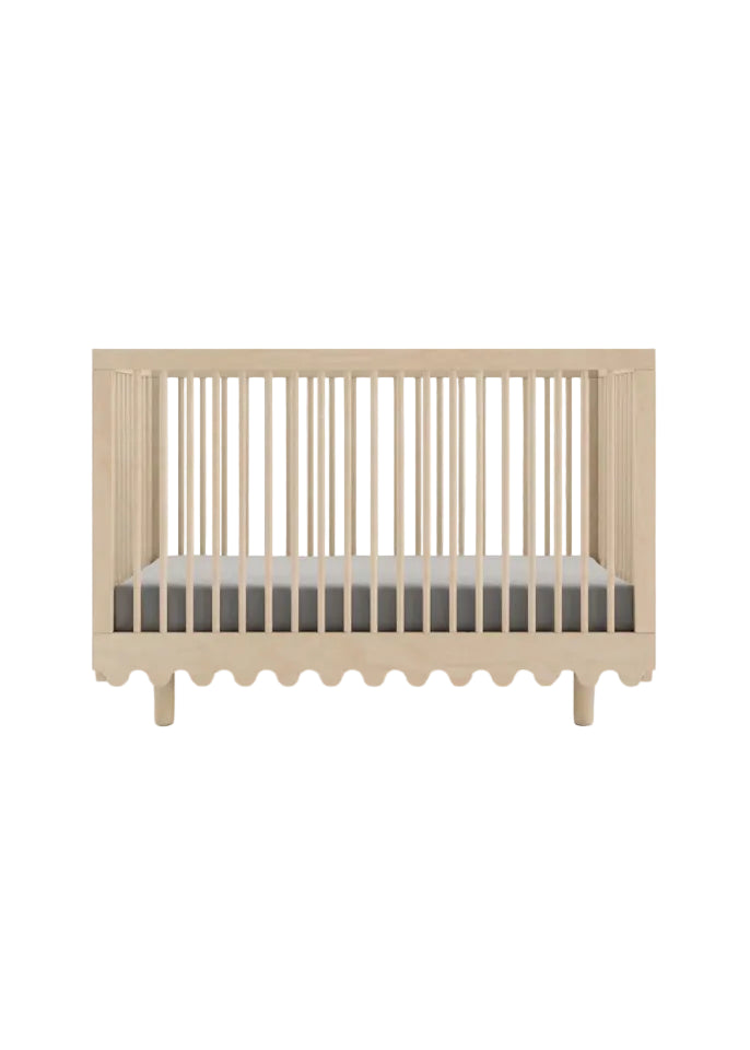 A light natural wood baby crib with vertical slats and a scalloped bottom edge, shown with a mattress inside on a clean white background.