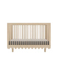 A light natural wood baby crib with vertical slats and a scalloped bottom edge, shown with a mattress inside on a clean white background.