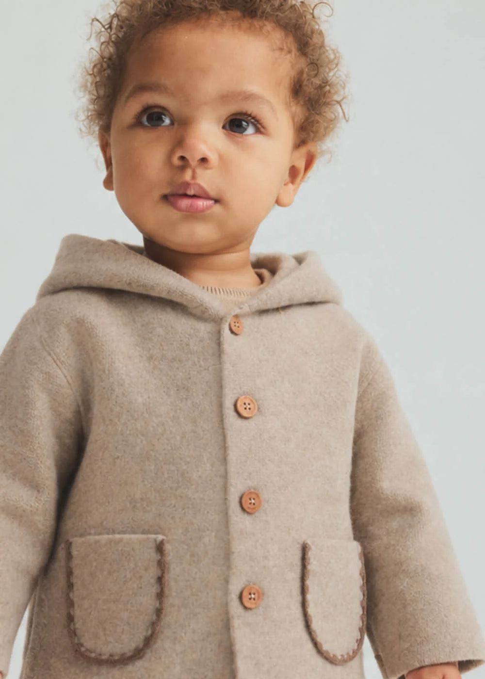 A close-up of a toddler wearing a light beige hooded cardigan with large front pockets and wooden buttons, photographed against a soft light gray background.