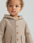 A close-up of a toddler wearing a light beige hooded cardigan with large front pockets and wooden buttons, photographed against a soft light gray background.