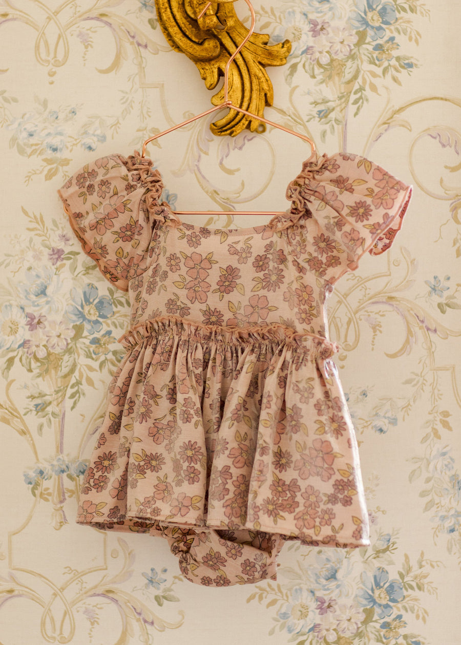 Floral dress with ruffled sleeves and bottom hanging on a decorative gold hanger against a floral wallpaper background.