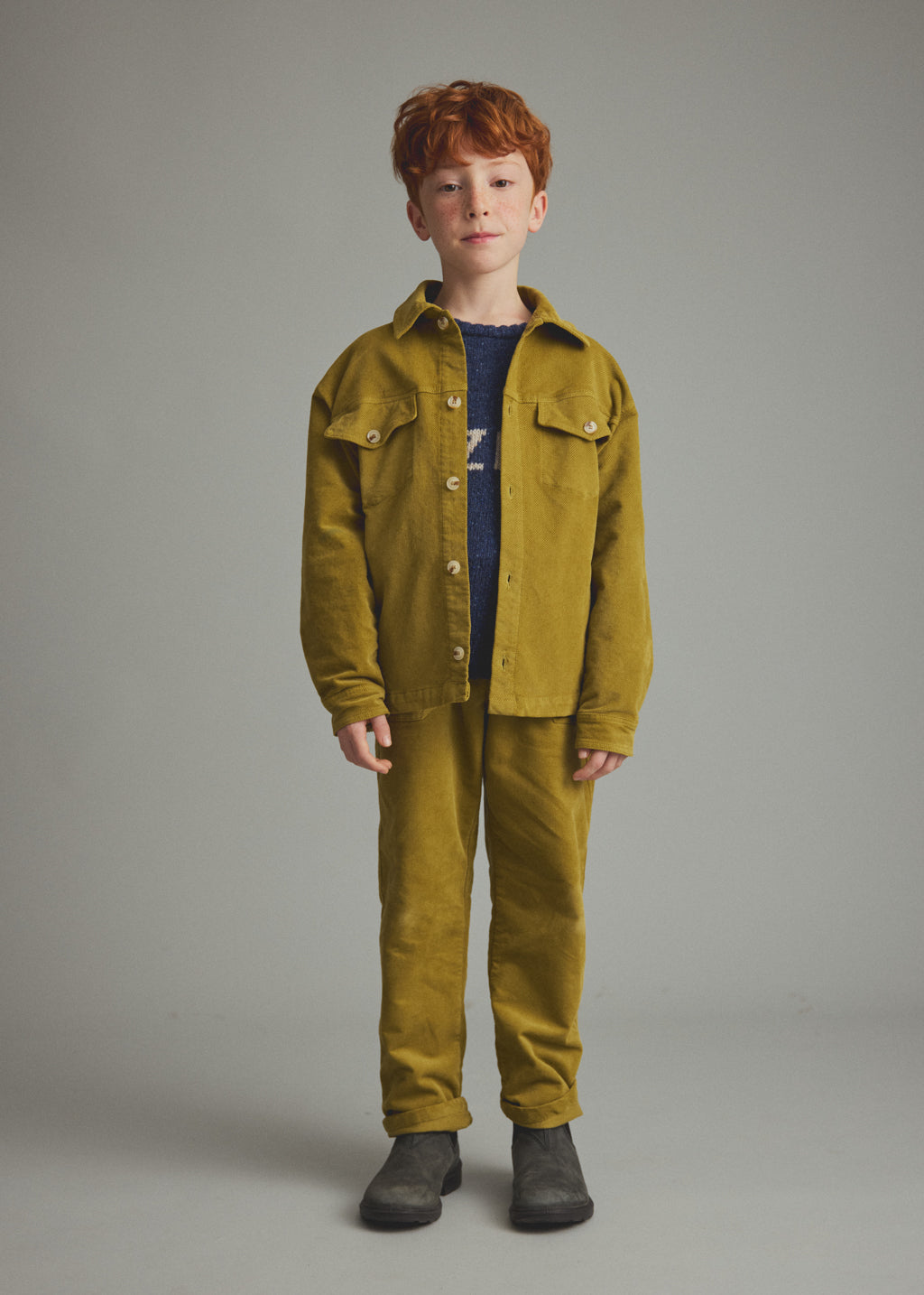 A young boy with red hair stands against a neutral gray studio background wearing a matching mustard yellow corduroy jacket and trousers set, a navy blue t-shirt, and dark gray boots.