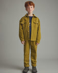A young boy with red hair stands against a neutral gray studio background wearing a matching mustard yellow corduroy jacket and trousers set, a navy blue t-shirt, and dark gray boots.