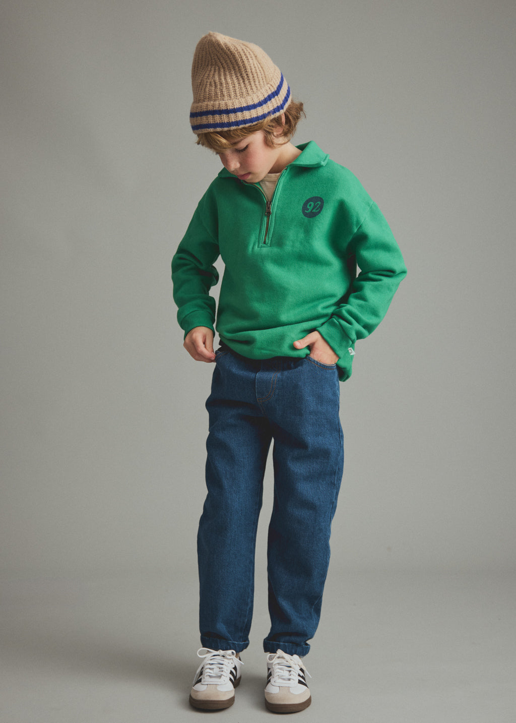 A child wearing a bright green half-zip pullover, blue jeans, white sneakers, and a beige knit beanie with blue stripes, standing against a plain gray studio background.
