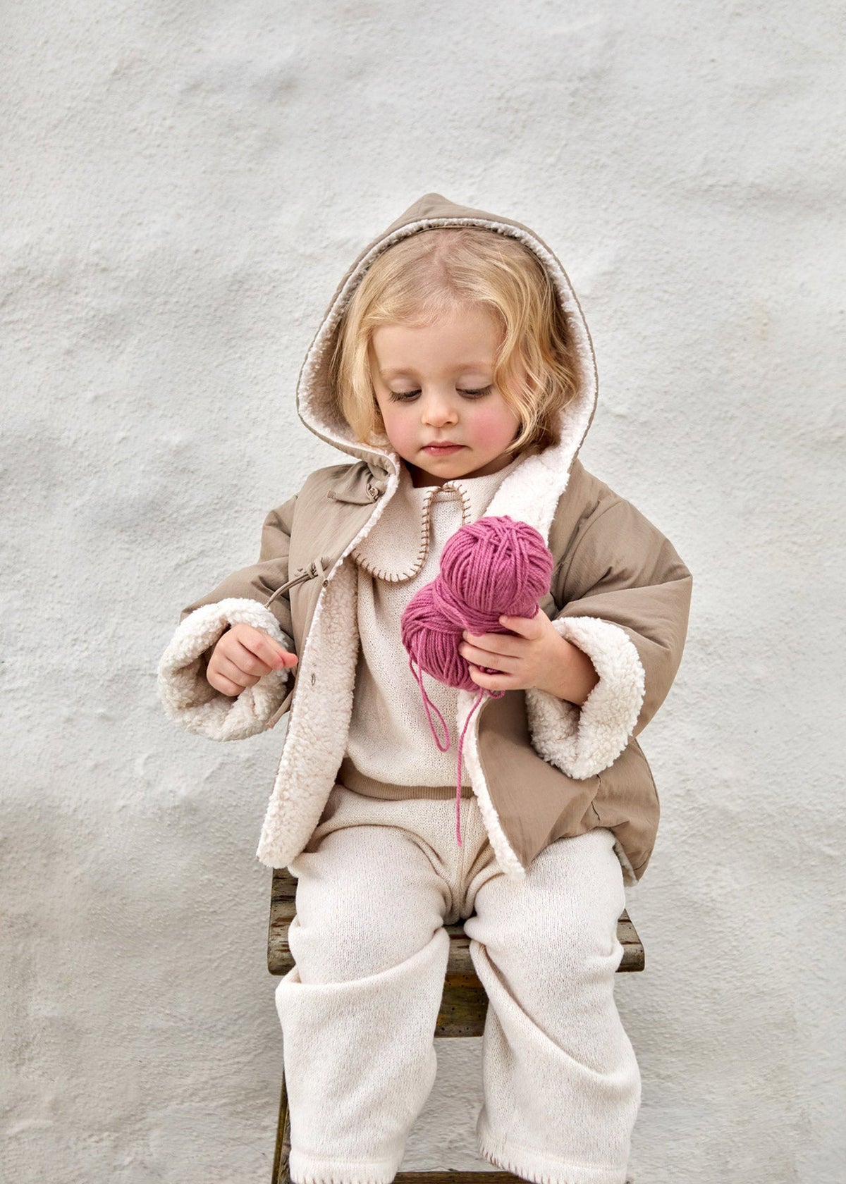 A young child wearing a beige hooded jacket with cream sherpa lining and cream pants, sitting on a wooden stool against a textured light gray wall and holding a ball of pink yarn.