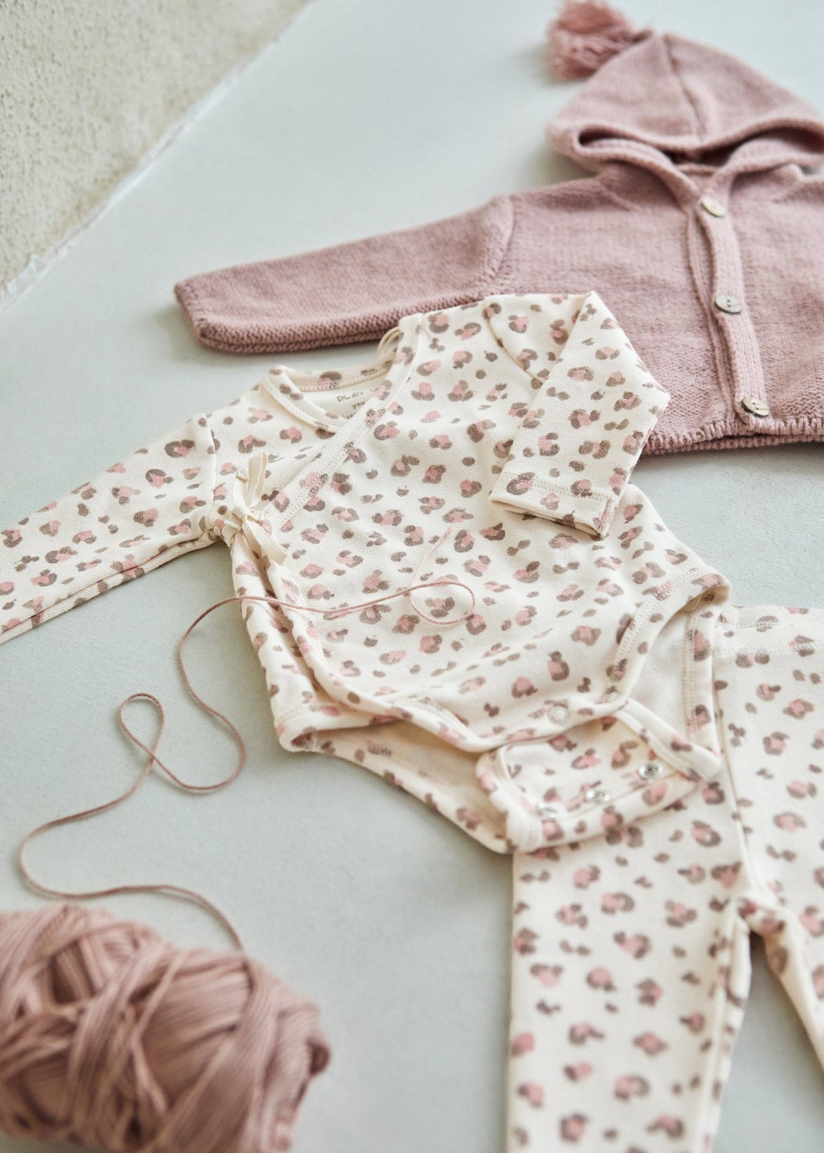 A baby clothing set laid flat on a light surface, including a cream bodysuit with a pink animal print, matching pants, and a dusty pink hooded cardigan. A ball of matching pink yarn sits in the lower left corner.