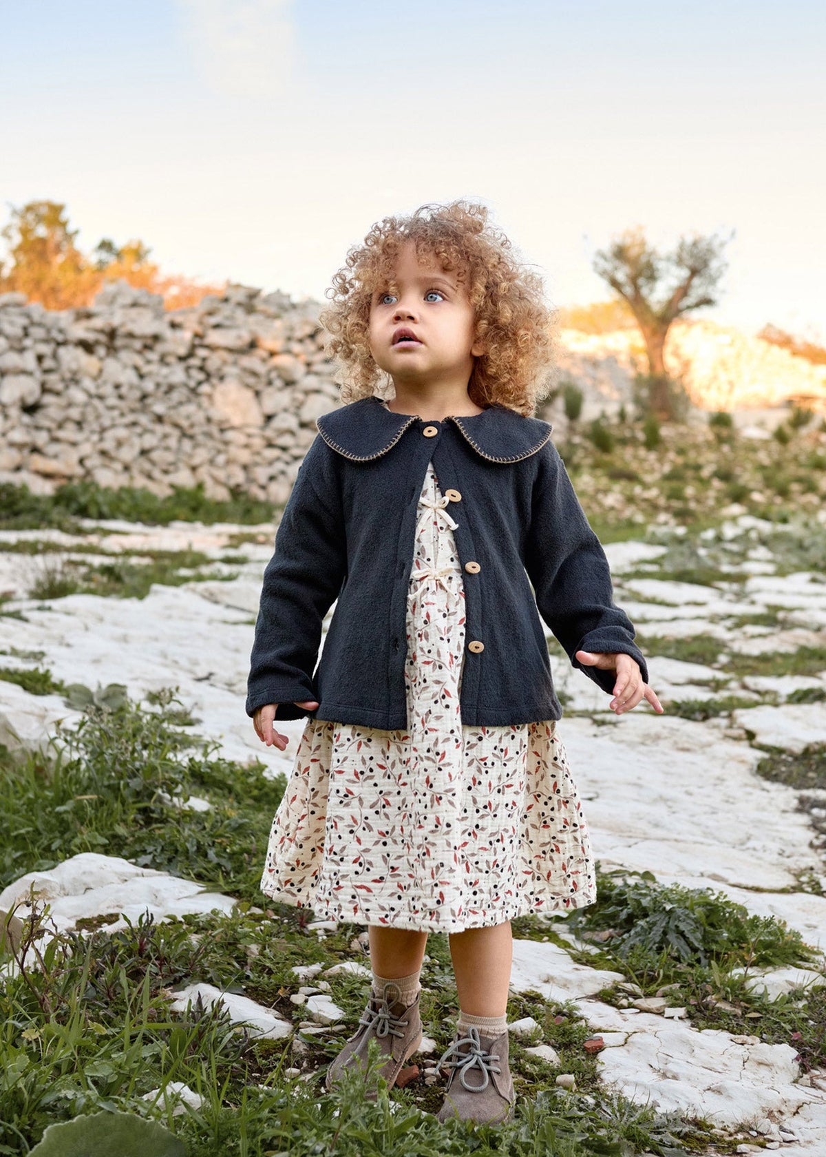 A young child with curly light brown hair stands outdoors wearing a dark navy cardigan over a cream floral dress, paired with brown boots. The background features a rustic stone wall and dry grass under a warm sky.