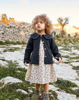 A young child with curly light brown hair stands outdoors wearing a dark navy cardigan over a cream floral dress, paired with brown boots. The background features a rustic stone wall and dry grass under a warm sky.