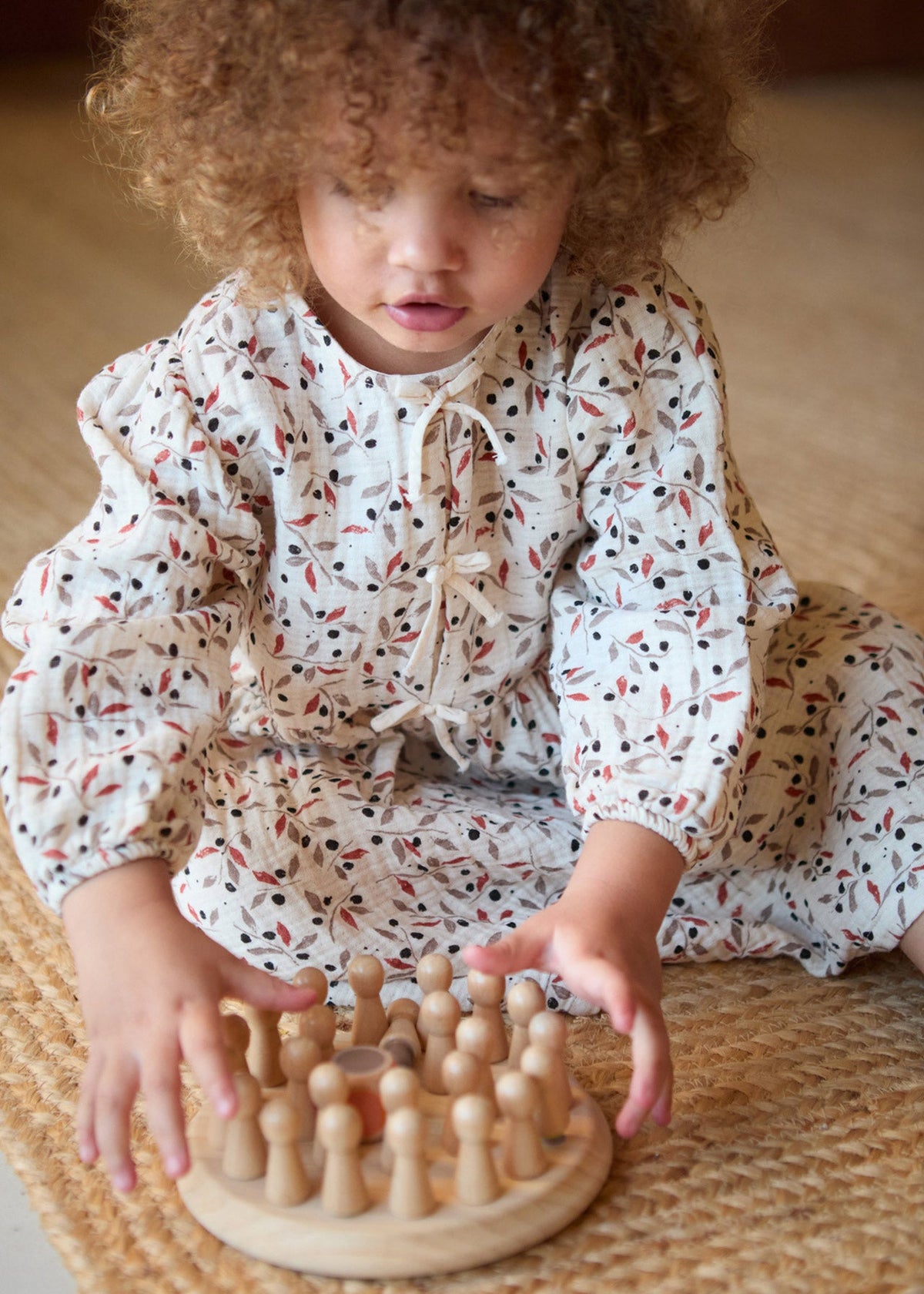 A toddler with curly hair sits on a woven mat, wearing a white long-sleeve romper with a red and black leaf pattern, interacting with a wooden peg board toy.
