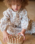 A toddler with curly hair sits on a woven mat, wearing a white long-sleeve romper with a red and black leaf pattern, interacting with a wooden peg board toy.