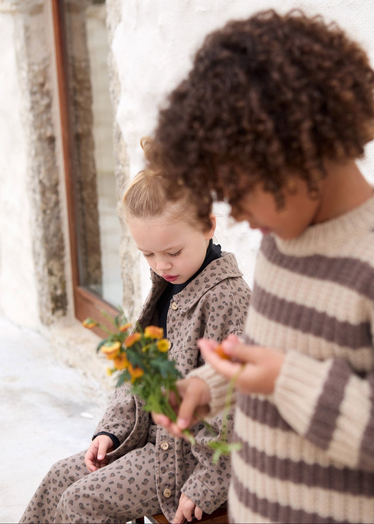 Two children outdoors near a stone wall, one wearing a beige and brown spotted pajama set and holding small yellow flowers, while the other wears a cream and brown striped knit sweater.