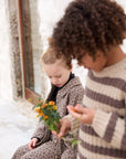 Two children outdoors near a stone wall, one wearing a beige and brown spotted pajama set and holding small yellow flowers, while the other wears a cream and brown striped knit sweater.