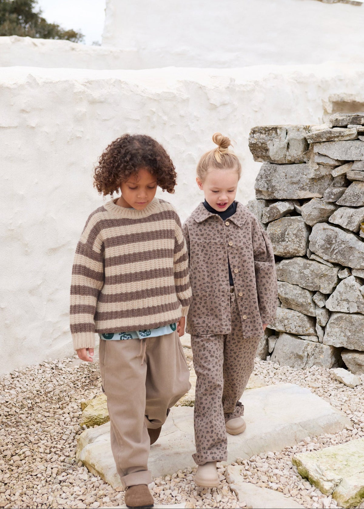 Two young children walking outdoors on a stone path in front of a white stucco wall and rustic stone wall. The child on the left wears a brown and beige striped knit sweater and beige pants, while the child on the right wears a matching brown patterned jacket and pants set.