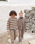 Two young children walking outdoors on a stone path in front of a white stucco wall and rustic stone wall. The child on the left wears a brown and beige striped knit sweater and beige pants, while the child on the right wears a matching brown patterned jacket and pants set.