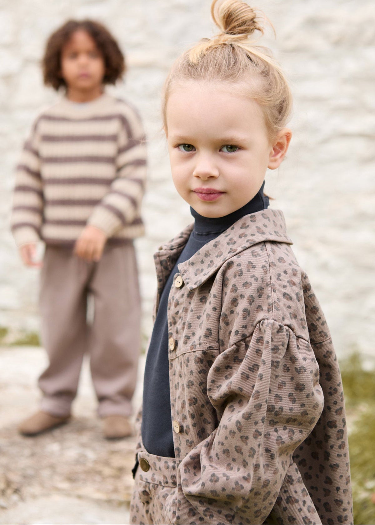 A young girl in the foreground wears a brown leopard-print jacket over a dark navy turtleneck, while another child stands blurred in the background wearing a beige and brown striped sweater.