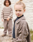 A young girl in the foreground wears a brown leopard-print jacket over a dark navy turtleneck, while another child stands blurred in the background wearing a beige and brown striped sweater.