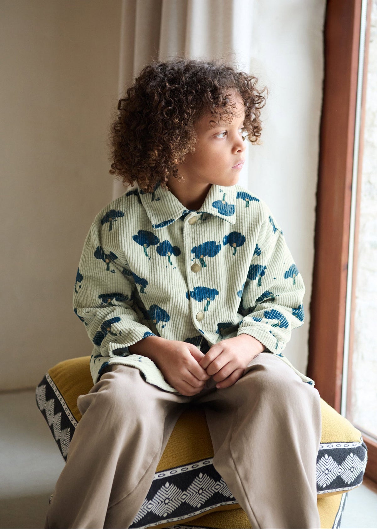 A child with curly brown hair sits on a yellow cushioned stool by a window, wearing a light sage green corduroy shirt with a blue tree pattern and beige pants.