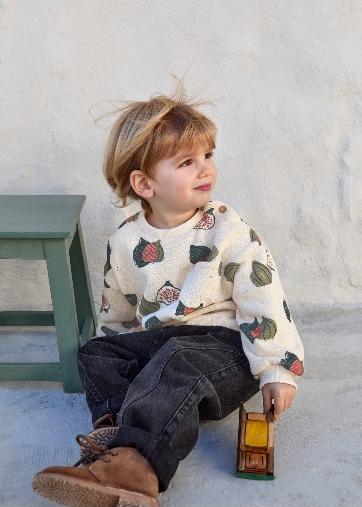 A toddler sitting on a concrete floor against a textured white wall, wearing a cream-colored crewneck sweatshirt with a colorful strawberry print, dark jeans, and brown boots, holding a small wooden toy truck.