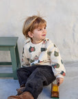 A toddler sitting on a concrete floor against a textured white wall, wearing a cream-colored crewneck sweatshirt with a colorful strawberry print, dark jeans, and brown boots, holding a small wooden toy truck.