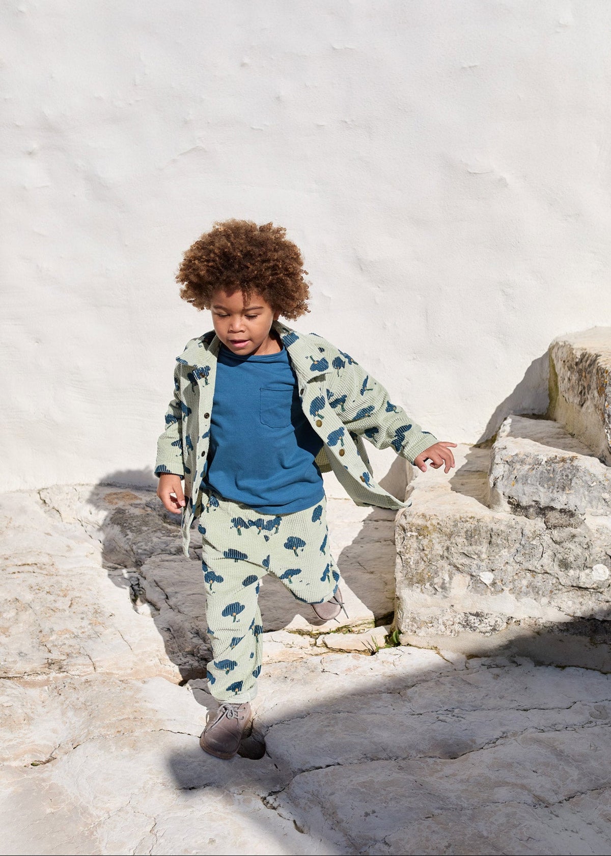 A young child with curly brown hair walks down light-colored stone steps against a white stucco wall, wearing a matching light green and dark green dinosaur print jacket and pants set over a blue t-shirt.