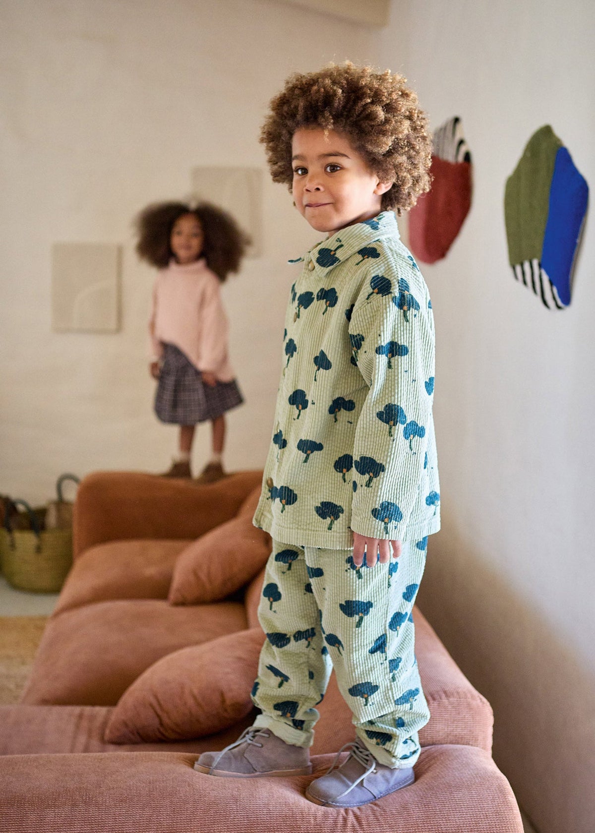 A young child with curly hair stands on a pink sofa wearing a light green two-piece pajama set with a blue cloud pattern. Another child is visible in the background of a living room setting.