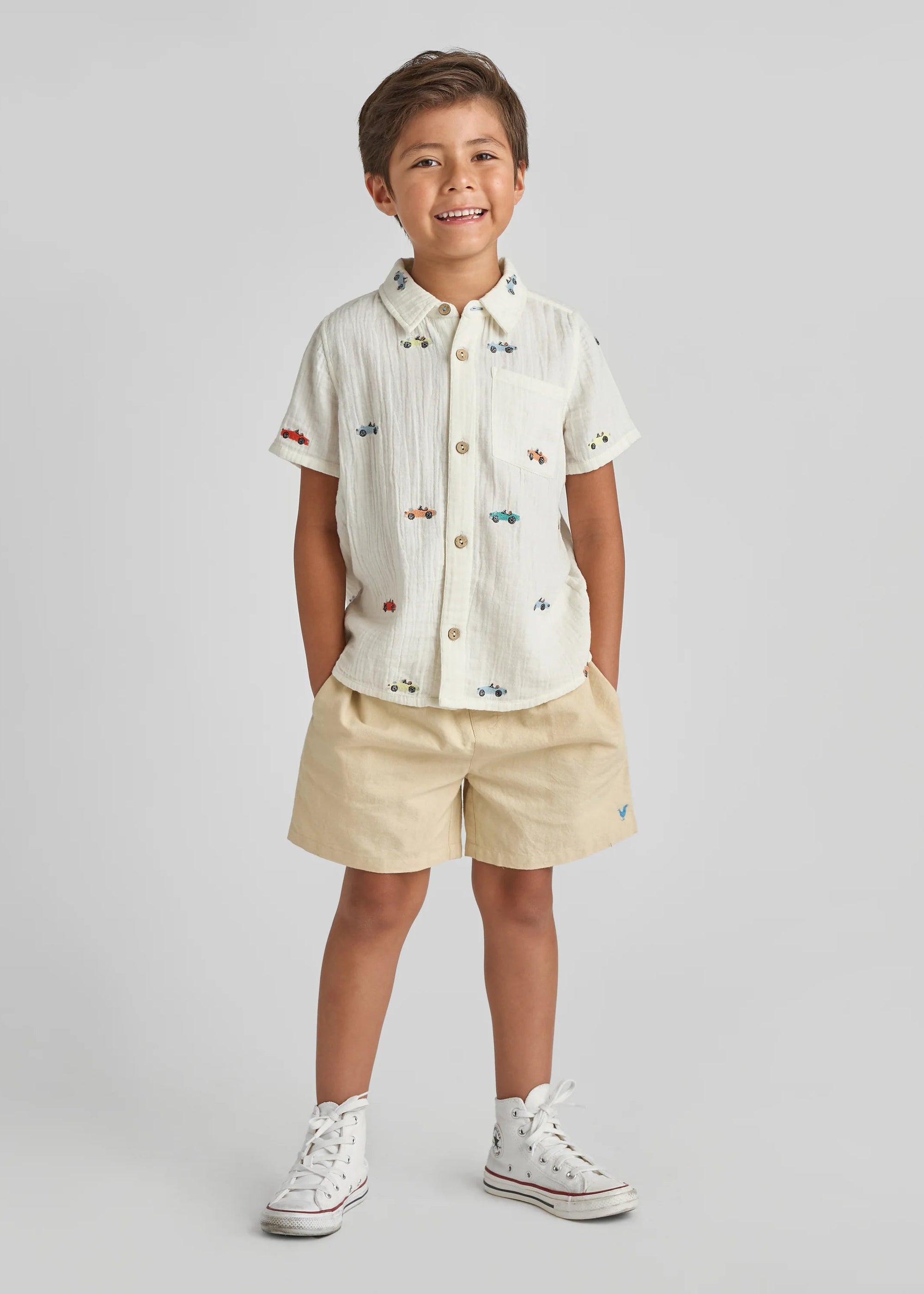 Child wearing a white shirt with colorful buttons and beige shorts on a light gray background